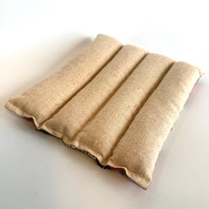 May include: A beige, rectangular heating pad with four long, parallel sections. The fabric has a textured appearance. The edges of the pad have a patterned border with red, black, and blue colours. The pad is designed for therapeutic use.