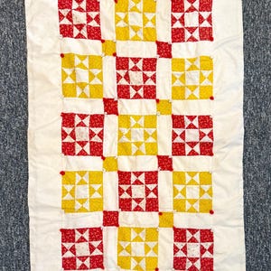 May include: A white quilt with a patchwork design of red and yellow squares. Each square is made up of smaller triangles, creating a star pattern. The quilt is edged with a white border.