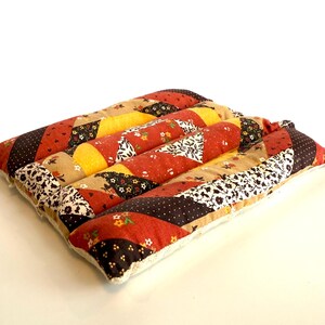 May include: A square, quilted cushion with a patchwork design. The cushion features strips of fabric in various patterns and colours, including red, yellow, brown, and floral prints. The edges are finished with a cream-coloured trim.