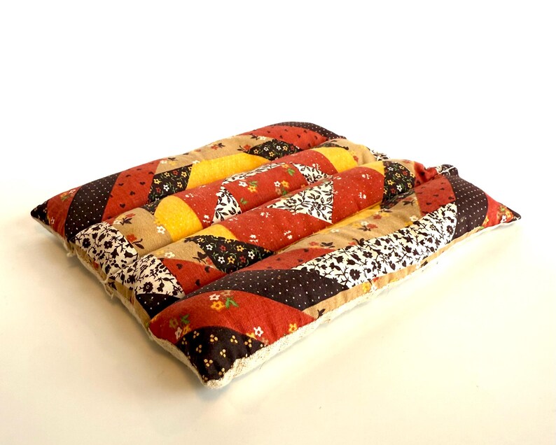 May include: A square, quilted cushion with a patchwork design. The cushion features strips of fabric in various patterns and colours, including red, yellow, brown, and floral prints. The edges are finished with a cream-coloured trim.