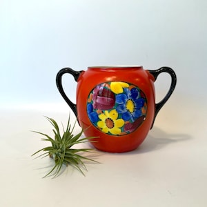 May include: A vintage orange ceramic sugar bowl with black handles and a floral design in shades of blue, yellow, and red. The bowl has a round shape and a white interior.