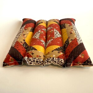 May include: Four rectangular, quilted fabric items with a patchwork design. The colour palette includes shades of red, brown, yellow, and beige. The fabric is patterned with floral and polka dot motifs. The edges are finished with a ruffled trim.