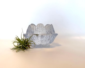 May include: A clear glass bowl with a textured, frosted finish. The bowl has a scalloped edge and a rounded shape. It is sitting on a white surface.