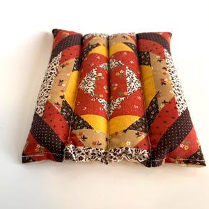 May include: A rectangular patchwork pillow in red, brown, yellow, and beige. The pillow is divided into four sections, each with a unique pattern, including floral and polka dot designs. The edges are finished with a floral trim. The pillow is made of fabric.