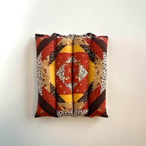 May include: A square, quilted cushion with a patchwork design. The cushion features a central diamond pattern in yellow and floral fabric, surrounded by red, brown, and beige geometric shapes. The edges are a mix of floral and polka dot patterns.