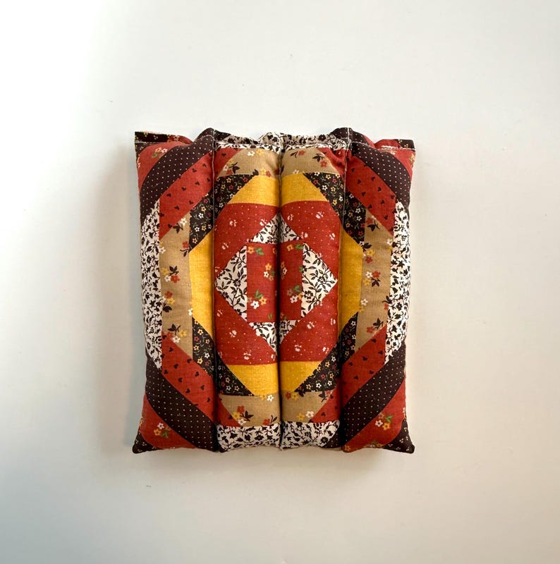 May include: A square, quilted cushion with a patchwork design. The cushion features a central diamond pattern in yellow and floral fabric, surrounded by red, brown, and beige geometric shapes. The edges are a mix of floral and polka dot patterns.
