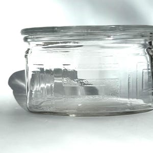 May include: A clear, rectangular glass jar with a wide mouth and a matching lid. The jar has a decorative, etched design on the front and sides. The jar is sitting on a white surface.