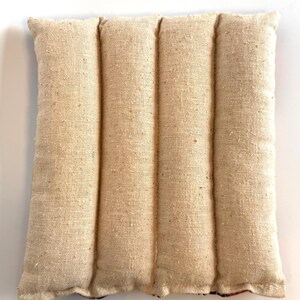 May include: A beige rectangular heating pad with four vertical sections. The pad is made from a textured, woven fabric, with visible seams. The bottom edge has a hint of patterned fabric. The pad is designed for therapeutic use.