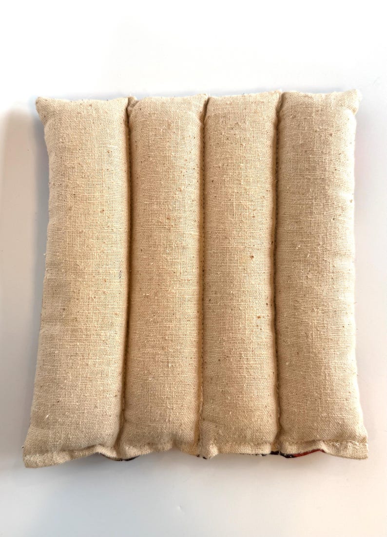 May include: A beige rectangular heating pad with four vertical sections. The pad is made from a textured, woven fabric, with visible seams. The bottom edge has a hint of patterned fabric. The pad is designed for therapeutic use.