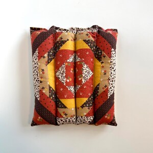 May include: A patchwork cushion with a quilted design. The cushion has a central square pattern in red, yellow, and white floral fabric, framed by diagonal stripes in shades of red, brown, and tan. The edges are finished with a floral print.