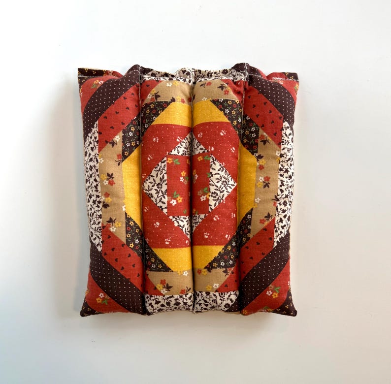 May include: A patchwork cushion with a quilted design. The cushion has a central square pattern in red, yellow, and white floral fabric, framed by diagonal stripes in shades of red, brown, and tan. The edges are finished with a floral print.