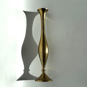May include: A tall, slender, gold-colored candlestick holder. The candlestick has a flared top and base, with a long, elegant stem. The candlestick is casting a shadow on a white surface.