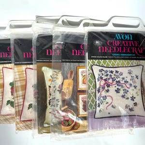 May include: A collection of Avon Creative Needlecraft crewel embroidery kits. The kits are packaged in clear plastic with white handles. The designs include floral and fruit-themed pillow patterns. The text on the packaging reads "Spring Violets Pillow".