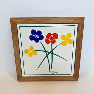 May include: A framed ceramic tile featuring a floral design. The artwork showcases three flowers in blue, red, and yellow, with green stems against a white background. The tile is set within a light brown wooden frame. The word "Salteras" is visible.