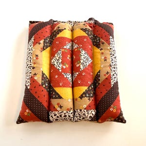 May include: A rectangular, quilted pillow with a patchwork design. The pillow features a combination of red, yellow, brown, and beige fabrics with floral and polka dot patterns. The pillow is made of four cylindrical sections.