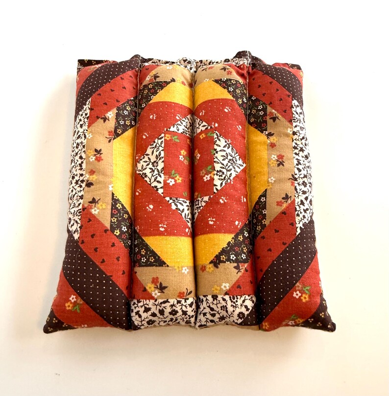 May include: A rectangular, quilted pillow with a patchwork design. The pillow features a combination of red, yellow, brown, and beige fabrics with floral and polka dot patterns. The pillow is made of four cylindrical sections.