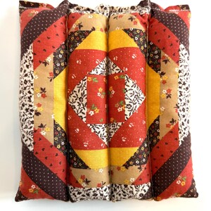 May include: A quilted pillow with a patchwork design. The pillow features a central diamond pattern in red, yellow, and floral fabric, surrounded by diagonal stripes in brown, red, and floral prints. The edges are finished with a floral fabric.