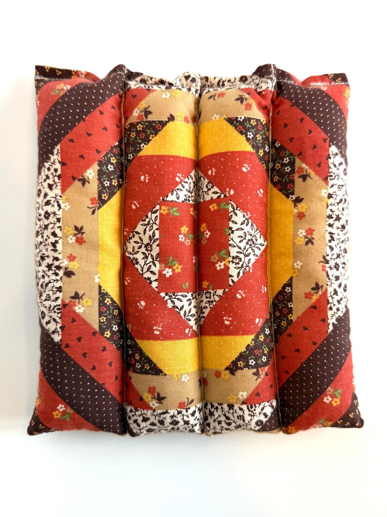 May include: A quilted pillow with a patchwork design. The pillow features a central diamond pattern in red, yellow, and floral fabric, surrounded by diagonal stripes in brown, red, and floral prints. The edges are finished with a floral fabric.