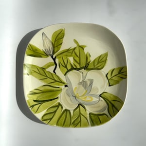 May include: Square ceramic plate with a hand-painted magnolia design. The plate features a large white magnolia flower with yellow accents, surrounded by green leaves and a bud. The background is a creamy off-white.