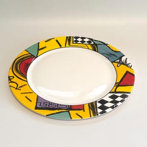 May include: Oval serving platter with a white center and a vibrant, patterned border. The border features yellow, red, black, and teal geometric shapes, including a checkerboard design. A decorative piece for serving food.