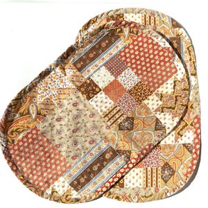 May include: Oval-shaped quilted placemats with a patchwork design. The placemats feature a mix of floral, paisley, and geometric patterns in shades of brown, beige, orange, and red. The edges are finished with a brown trim.
