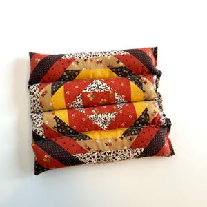 May include: Four stacked rectangular pillows with a patchwork design. The pillows feature red, yellow, brown, and floral patterned fabrics. The design includes diagonal stripes and diamond shapes, creating a vintage, quilted aesthetic.