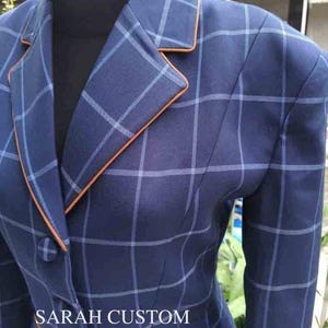 Op de afbeelding: Een marineblauwe blazer met een ruitpatroon en oranje bies. De blazer heeft een revers en een sluiting met één knoop. De tekst "SARAH CUSTOM STORE IN" is zichtbaar onderaan de afbeelding.