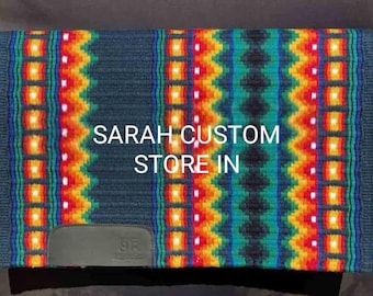 Pode incluir: Uma almofada de sela de cavalo colorida com um padrão geométrico em tons de azul, laranja, amarelo e verde. A almofada tem um remendo de couro com as iniciais "SR" e o texto "SARAH CUSTOM STORE IN" em branco.