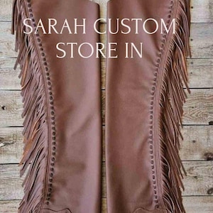May include: Brown leather chaps with decorative floral tooling and fringe detailing. The chaps feature a belt with a silver buckle and the text "SARAH CUSTOM STORE IN". The chaps are designed for riding and western wear.