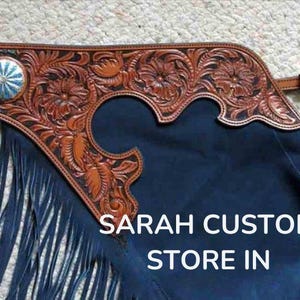 May include: Dark blue leather chaps with detailed floral tooling in brown. The chaps have a silver concho, fringe, and a buckle with turquoise detailing. The text "SARAH CUSTOM STORE IN" is visible.
