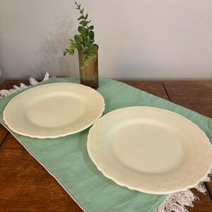 May include: Two cream-coloured ceramic plates with a floral embossed design on the rim. The plates are placed on a light green placemat with a fringed edge. A brown glass vase with greenery is in the background.