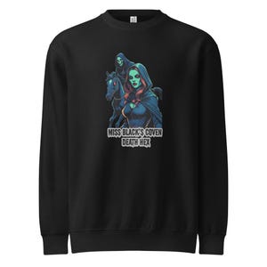 以下が含まれることがあります： 黒いスウェットシャツ。赤い髪とフード付きマントを着た女性が馬に乗っているグラフィックで、彼女の後ろには2人目の人物がいます。「MISS BLACK'S COVEN DEATH HEX」というテキストが画像の下にあります。