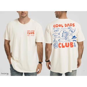 Pode incluir: T-shirts cor marfim com o texto "COOL DADS CLUB Est. 2026" em vermelho e azul. A frente da camisa tem o texto no peito esquerdo. A parte de trás da camisa tem um gráfico com o texto "COOL DADS CLUB" e outras ilustrações.