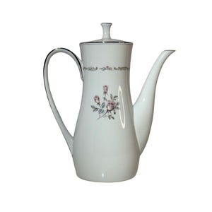 May include: White porcelain coffee pot with a silver trim and a floral rose design. The pot has a curved handle and a long spout. The lid has a small knob. The floral design features pink roses and green leaves.