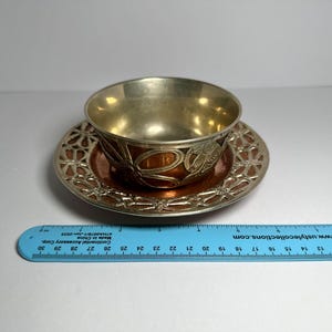 May include: A vintage teacup and saucer set. The cup and saucer have a metallic finish with a floral design. The saucer has an openwork design around the edge. The cup is approximately 2 inches tall.