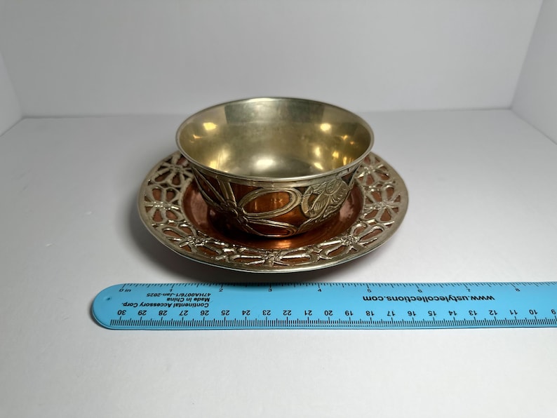 May include: A vintage teacup and saucer set. The cup and saucer have a metallic finish with a floral design. The saucer has an openwork design around the edge. The cup is approximately 2 inches tall.