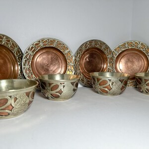 May include: A set of decorative metal plates and bowls. The plates have a copper center with a silver filigree border. The bowls have a silver exterior with copper and floral designs. The set includes four plates and three bowls.