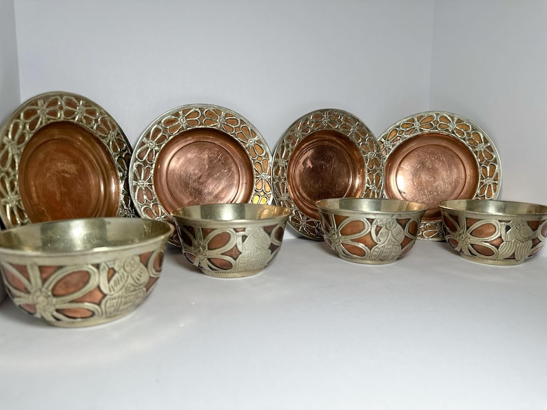 May include: A set of decorative metal plates and bowls. The plates have a copper center with a silver filigree border. The bowls have a silver exterior with copper and floral designs. The set includes four plates and three bowls.