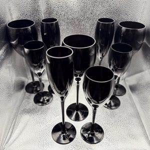 May include: An assortment of black stemware, including wine glasses and champagne flutes, displayed on a reflective surface. The glasses have a sleek, modern design with a dark, opaque finish, creating a sophisticated aesthetic. The set includes various sizes and shapes.
