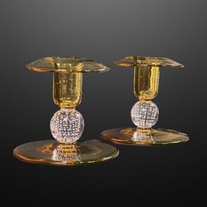 May include: Two amber glass candle holders. Each has a round base, a clear glass sphere, and a flared top. The candle holders are made of glass and have a reflective surface. The background is dark.