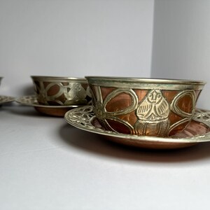 May include: A set of three copper and silver-toned teacups and saucers. The cups have a copper base with silver-toned rims and decorative patterns. The saucers are silver-toned with an openwork design. The teacups and saucers are likely vintage.
