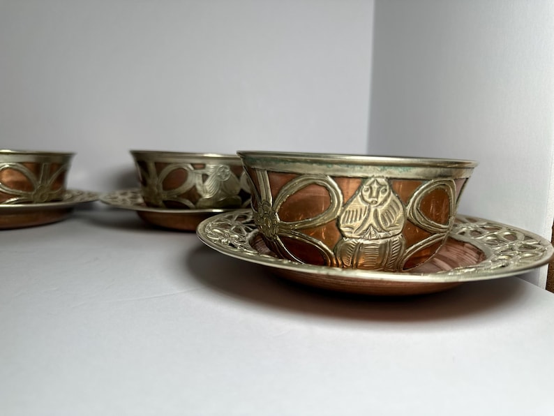 May include: A set of three copper and silver-toned teacups and saucers. The cups have a copper base with silver-toned rims and decorative patterns. The saucers are silver-toned with an openwork design. The teacups and saucers are likely vintage.