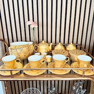 May include: A golden tea set with a floral design, including teacups, saucers, a teapot, sugar bowls, and serving bowls. The set is arranged on a gold-colored serving cart, with a vase of pink flowers in the background.
