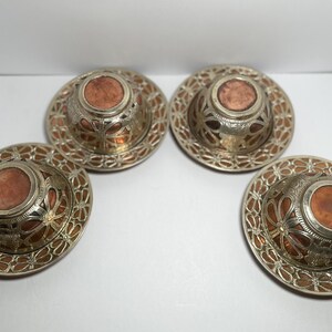 May include: Four ornate metal coasters with a copper-colored center and intricate silver filigree design. The coasters have a circular shape and are arranged in a square formation, showcasing their decorative patterns.