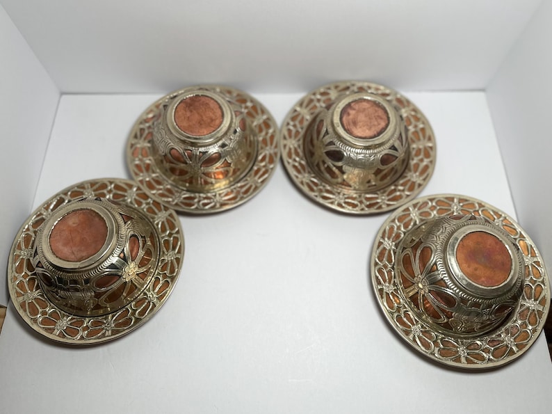 May include: Four ornate metal coasters with a copper-colored center and intricate silver filigree design. The coasters have a circular shape and are arranged in a square formation, showcasing their decorative patterns.