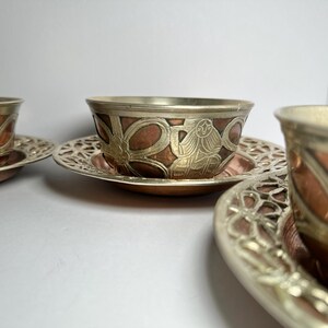 May include: Set of three decorative metal teacups and saucers. The cups have a copper-colored interior and are adorned with silver-colored metal filigree and detailed designs. The saucers feature a matching openwork pattern.