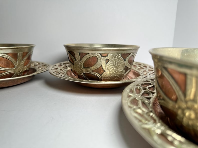 May include: Set of three decorative metal teacups and saucers. The cups have a copper-colored interior and are adorned with silver-colored metal filigree and detailed designs. The saucers feature a matching openwork pattern.