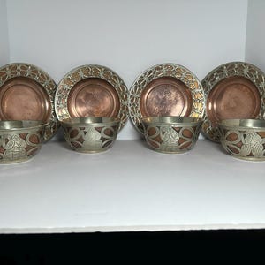 May include: A set of four copper-colored plates and four matching bowls. The plates have a decorative silver-colored border. The bowls have a silver-colored exterior with a copper-colored interior and decorative design.