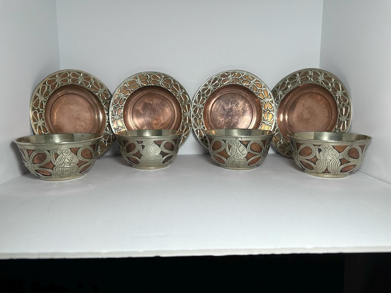 May include: A set of four copper-colored plates and four matching bowls. The plates have a decorative silver-colored border. The bowls have a silver-colored exterior with a copper-colored interior and decorative design.