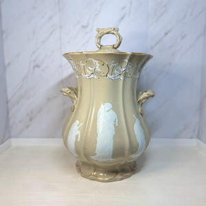May include: A beige ceramic vase with a lid, featuring white relief figures and decorative accents. The vase has a gold rim and handles, and a loop handle on the lid. The design is reminiscent of classical art.